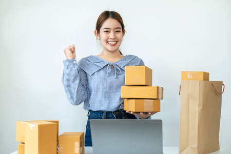 Asian woman small business owner standing and smiling to raise arm when job finished after receive order from customer and packing product to parcel of box at home office for delivery to clientの写真素材