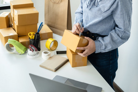 Asian woman small business owner standing to writing customer address on parcel of box and checking information on laptop while packing product deliver to client at homeの写真素材