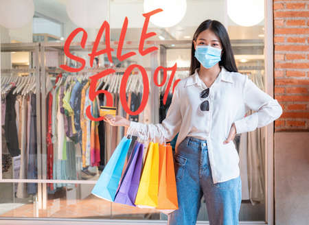 Asian woman shopaholic wearing face mask with many colorful shopping bags and holding credit card for payment in hand at shopping mallの写真素材