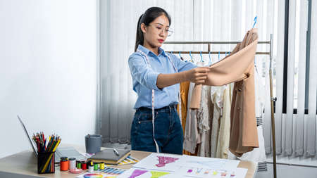 Asian woman fashion designer holding new clothes in hanger and looking on new clothes collection while standing to working in her workshopの写真素材