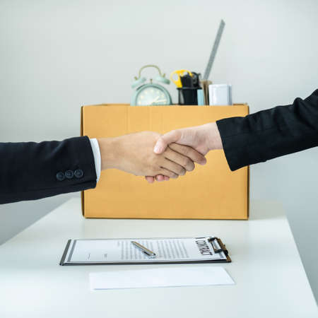 Close up of business woman employee in suit shaking hands with boss after signing on resignation letter and carrying packed personal belongings in brown cardboard box putting on the table in officeの写真素材