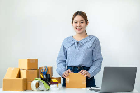 Asian woman small business owner smiling to looking at the camera and standing to holding parcel of box at the table while preparing to packing product deliver to client at homeの写真素材