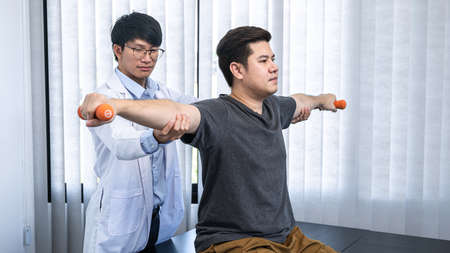 Young asian physiotherapist stretching the arms of man patient holding a dumbbell in hands during rehabilitation therapies and treatment of physiological disorders in clinicの写真素材