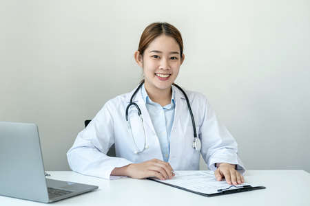 The doctor is diagnosing the patient's disease to writing symptom and a prescription on clipboard while smiling to looking at the camera and sitting to working in her clinicの写真素材