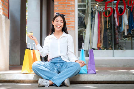 Asian shopaholic woman wearing sunglasses headband with many colorful shopping bags and holding credit card while sitting to rest after make purchase and payment heavy shopping in shopping mallの写真素材