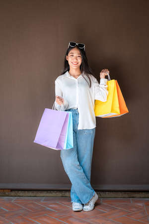 Asian shopaholic woman wearing sunglasses headband with many colorful shopping bags in both hands after heavy shopping isolated over brown backgroundの写真素材