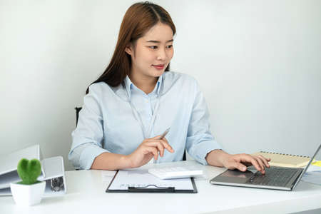 Woman office worker reading and typing business project report on laptop after that taking note of information on notebook while sitting to work hard for new business in officeの写真素材