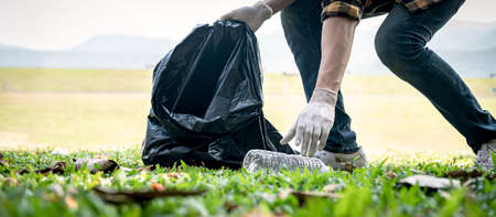 Volunteer man in gloves walking and stooping to collect plastic bottles into plastic black bag for cleaning the park during environmental activity to collecting garbageの写真素材