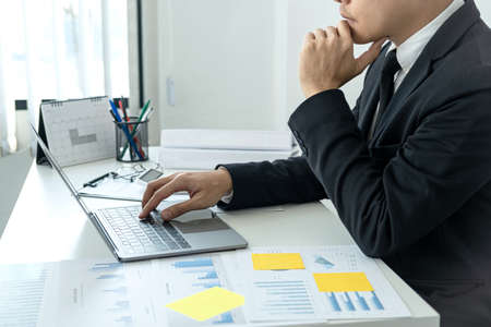 Businessman boss in suit is thinking and analysis of investments in new business from chart during to typing new idea for business project and financial data while sitting to working in officeの写真素材