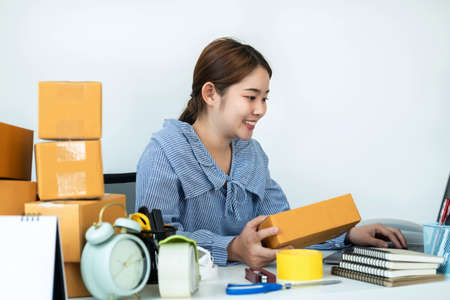 Asian woman small business owner holding box of parcel in hands and checking order on laptop while courier working with many boxes of parcel on the table to preparing delivery for customer at homeの写真素材