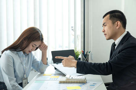 Businessman boss in suit scolding and pointing to woman office worker about bad work of her while she is sitting to stress and bow down in the officeの写真素材