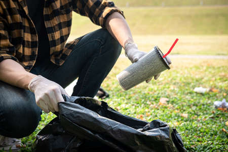 Volunteer man in gloves sitting to picking up coffee cup into plastic black bag for cleaning the park during environmental activity to collecting garbageの写真素材