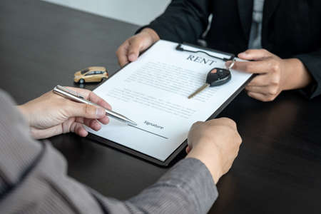 Asian salesman holding rental contract and hand over car key to client while he is holding pen to signing on the contract car trade or car loan or car rental on the table in the officeの写真素材