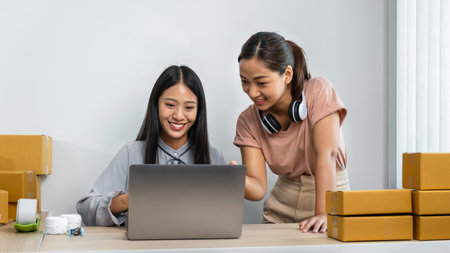 Two young business woman entrepreneur are working together with many parcel box and writing order on notebook, couriers are recheck address client on laptop and packing product sending home deliveryの写真素材