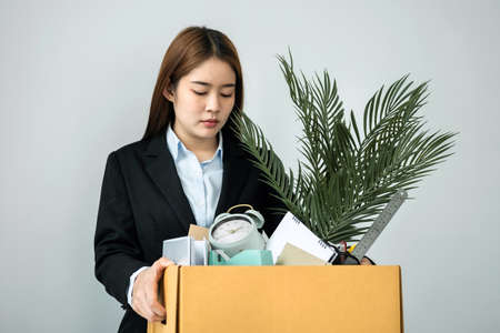 Business woman employee in suit stressful resignation from job while picking up personal belongings into brown cardboard box and carrying to walking out from officeの写真素材
