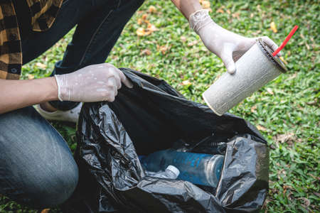Volunteer man in gloves sitting to picking up coffee cup into plastic black bag for cleaning the park during environmental activity to collecting garbageの写真素材