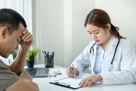 The patient is stressed and hand held forehead when listening disease and symptoms from doctor during she is writing diagnostic, prescription and to giving advice to him in her clinicの写真素材