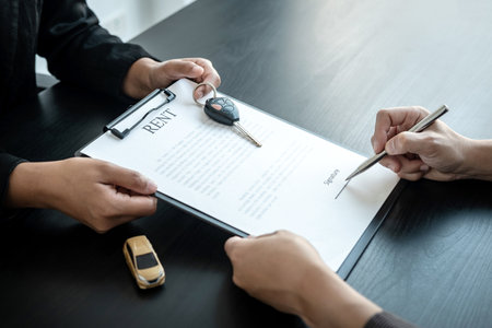 Asian salesman holding rental contract and hand over car key to client while he is holding pen to signing on the contract car trade or car loan or car rental on the table in the officeの写真素材