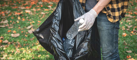 Volunteer man in gloves walking to carrying plastic black bag and collecting plastic bottles into...