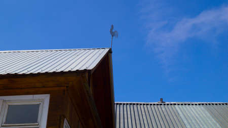 Stainless steel weather vane rooster on the roof of the house on the blue sky backgroundの写真素材