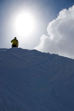 A man stands atop a snowy mountain ridge with sun and clouds behind.の写真素材