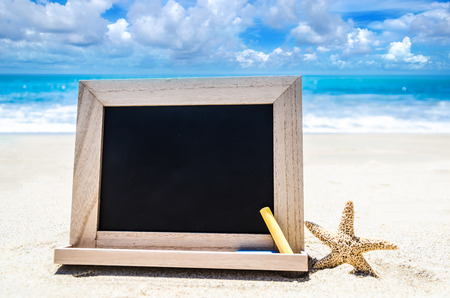Empty black wood chalkboard with chalk and starfish on the sandy beach near the oceanの写真素材