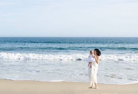 Mother is holding her adorable baby girl on the sandy beach near ocean in sunny day/Mothers day conceptの写真素材