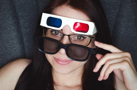 Smiling girl with 3 different kinds of glasses sits in the cinema and watches a movie through all of...