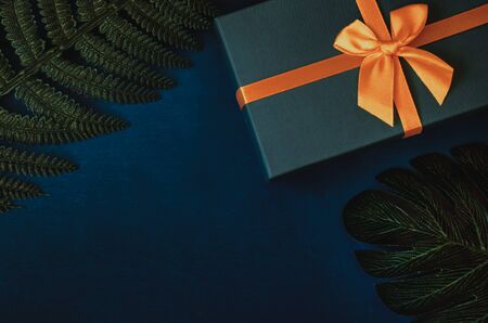 A gift box and leaves of fern and monstera on dark blue background flat lay top view. Stylish object arrangement for international men's day with copy spaceの写真素材
