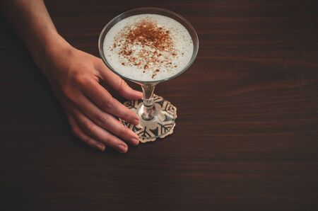 Female hand holds a glass of eggnog on wooden table background. Copy space