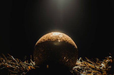 One golden round christmas toy on dark background close-up with volumetric light and deep shadows.