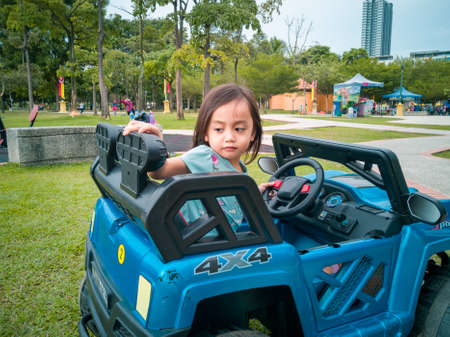 An Asian kid driving electric toy car in a tropical park. Outdoor toys. Children in battery power vehicle. Little girl riding toy truck in the garden. Family playing in the backyard.の写真素材