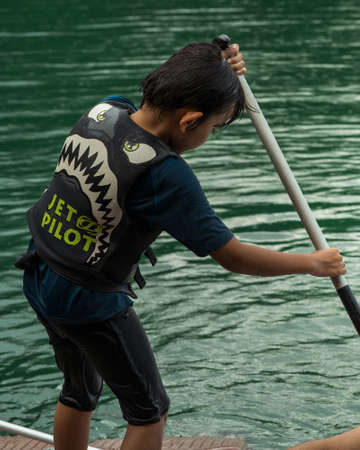 Kenyir, Malaysia - July 22, 2020: A boy wearing life jackets paddling on a boat. View from behind.のeditorial素材