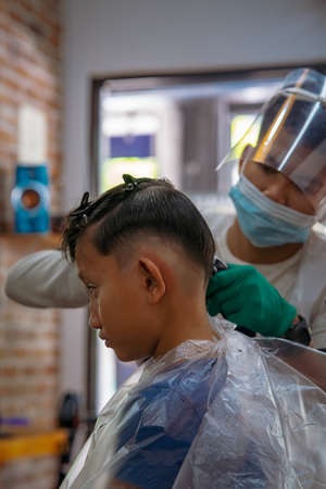 Children hairdresser with scissors is cutting little boy against in a barber shop.のeditorial素材