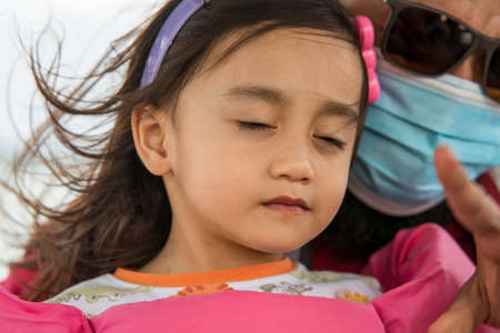 A portrait of toddler girl closing eyes feeling the wind while father in the background wearing face mask on the boat.の写真素材