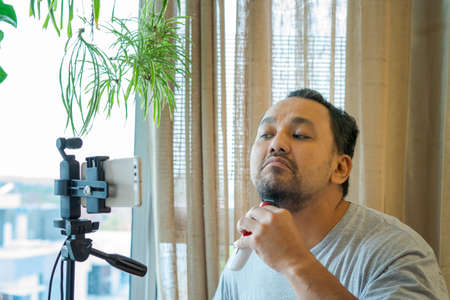 Asian man shaving beard with machine and recording the process using camera.
