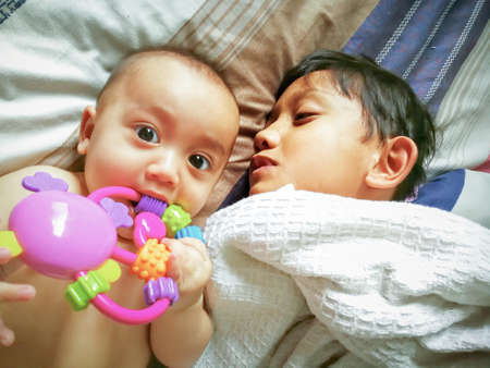 Portrait of sleepy brother on the bed with little baby sister playing with a toy