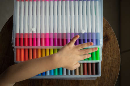 60 pieces of colorful markers in transparent plastic packaging on a wooden background. Top view with child hand.の写真素材