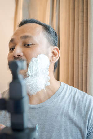 Shaving cream on the face of an Asian man. Man recording shaving process on the camera.の写真素材