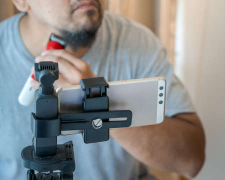 Camera recording with mobile phone of a man shaving beard with electric razor machine.の写真素材