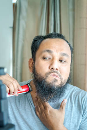 Matured Asian man shaving beard with electric razor machine.