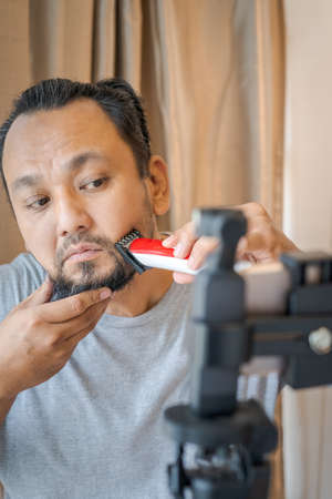 Asian man shaving beard with electric razor machine.
