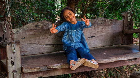 Little girl showing peace sign as sitting on the wooden swing in the garden.の写真素材