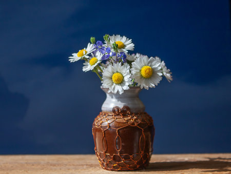 A delicate bouquet of daisies in a small vase. Still life with a bouquet of meadow flowers daisies in a decorative vase with a blue sky on a wooden background.の写真素材