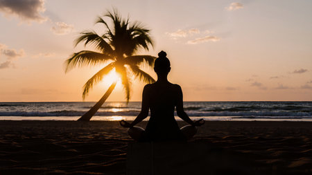 Meditating young woman practicing yoga at sunset. Yogi sitting in lotus position on sandy beach with palm tree at sea tide.の写真素材