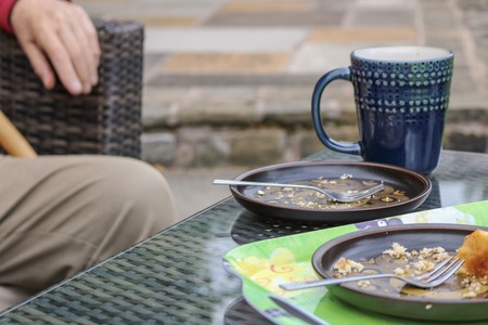 Blue and brown crockery with cake crumbs sat on a glass and wicker garden table with the leg and hand of a man in the shot.の写真素材