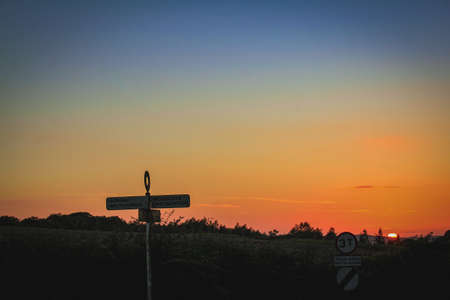 The sun sets behind fields, with a road sign in the foregroundの写真素材