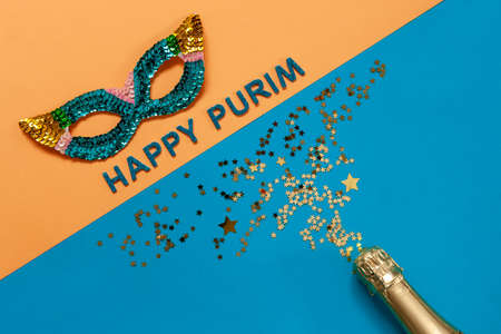 Carnival masquerade mask, champagne bottle and gold glitter confetti. Top view, Close up on blue and yellow colors background with Happy Purim text.の写真素材