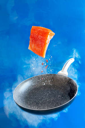 Close-up photo of fresh raw salmon, trout fillet floating in the air above the frying pan on the blue water backgroundの写真素材