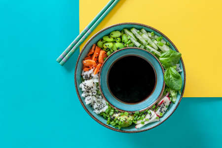 Pan Asian trendy Homemade Vegan Poke Bowl with Rice, edamame, fresh vegetables and soy sauce, served with chopsticks on vivid yellow and turquoise backgroundの写真素材